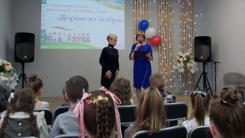 На сцене две женщины: одна в синем платье с микрофоном и планшетом, вторая в тёмном платье. Перед ними сидят дети. На экране сзади надпись «V юбилейный областной школьный фестиваль 2026 “Дорогою добра”», рядом гирлянда огней и связка шаров.