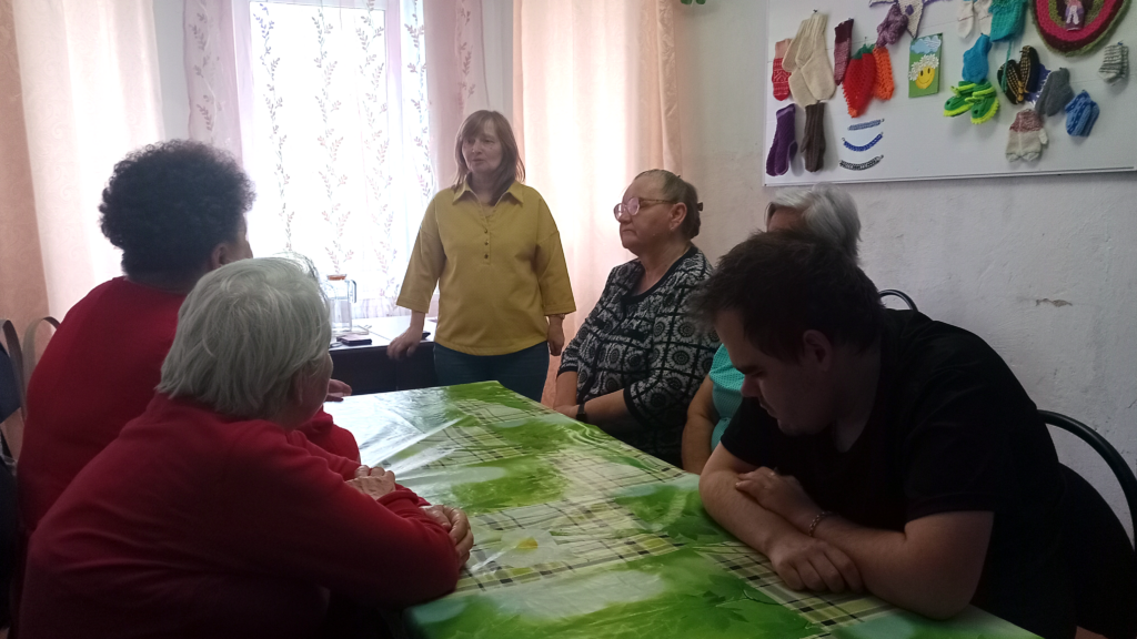 Люди сидят за столом с зелёной клеёнкой.
Спереди слева две пожилые женщины в красном, справа мужчина в чёрном, наклонился к столу.
Напротив стоит женщина в жёлтой блузке, рядом ещё две пожилые женщины.