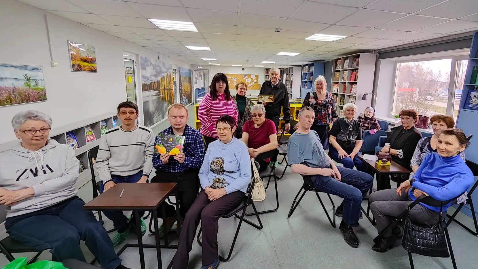 Группа из примерно 16 взрослых людей сидит и стоит в помещении библиотеки, смотрят в сторону камеры. В первом ряду мужчина держит открытку или коробку с изображением апельсинов. Вокруг – стеллажи с книгами и большие окна.