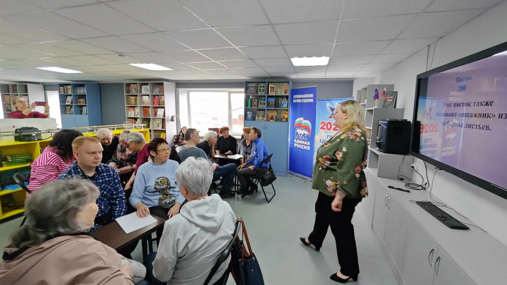 В библиотеке проходит мероприятие: люди сидят группами за столами, а женщина у большого экрана ведёт игру или викторину. На экране текст вопроса про цветок, на фоне видны стеллажи с книгами и стенды с логотипом «Единая Россия»