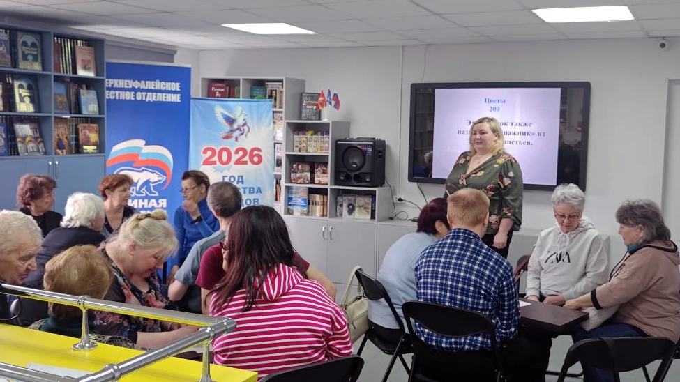 В библиотеке идёт викторина: люди сидят группами за столами, а женщина у экрана зачитывает вопрос про цветок. На заднем плане баннеры «Единая Россия» и «2026 – год семьи».