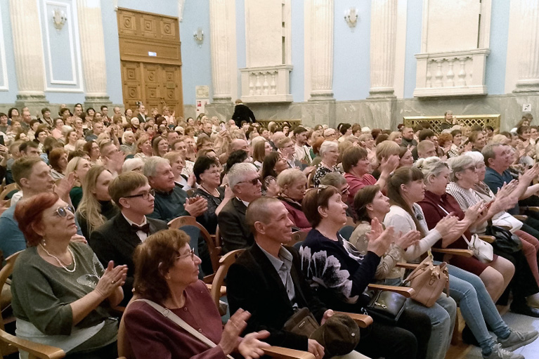 Большой зрительный зал, все ряды плотно заняты людьми.
Публика сидит на стульях с подлокотниками и дружно аплодирует в сторону сцены.