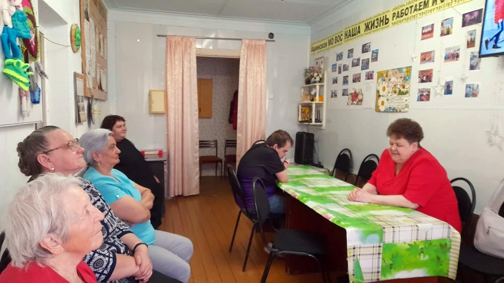 Небольшая комната, вдоль стола с зелёной клеёнкой сидят пожилые люди.
Слева в ряд несколько женщин, ближе к правому краю мужчина в чёрном, а напротив него женщина в ярко‑красной блузке.
На стене справа фотографии и надпись на жёлтой ленте: «НАША ЖИЗНЬ РАБОТАЕМ И ОТДЫХАЕМ».
Сзади по центру дверной проём с розовыми шторами.