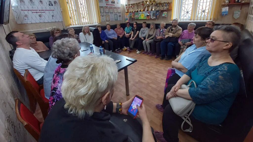 Люди сидят по периметру на стульях и диванах, внимательно слушают. На столе в центре стоят бутылки воды. На стенах — стенды, грамоты, наверху много кубков. На переднем плане женщина держит в руках включённый смартфон.
