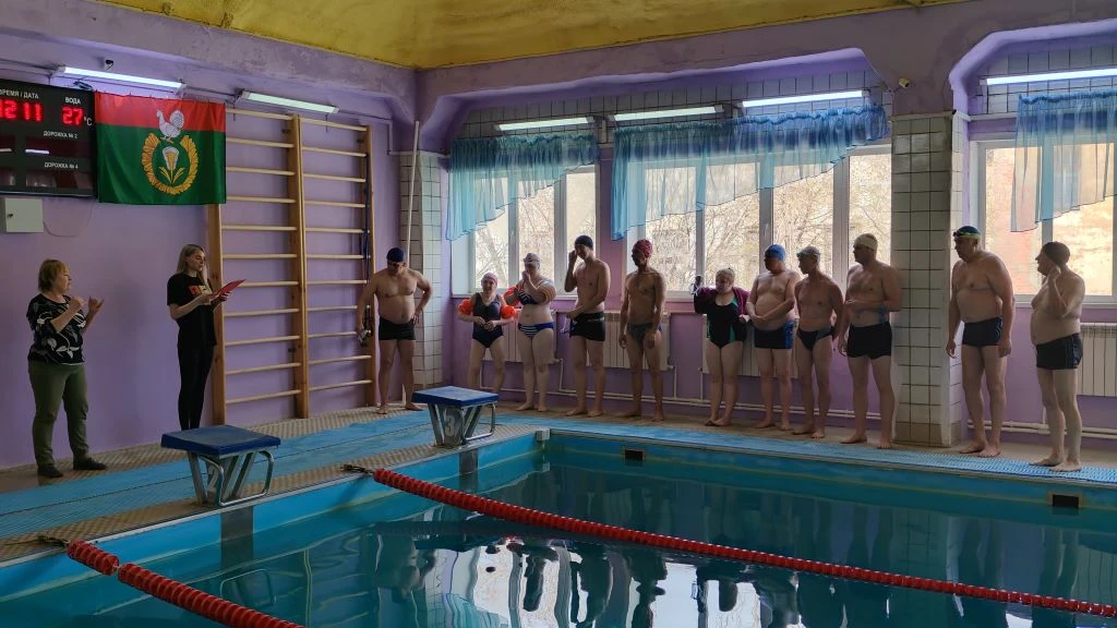 Закрытый бассейн. У бортика в ряд стоят участники в купальниках и шапочках, готовятся к заплыву. Слева две женщины, одна читает по листу. Над ними табло и зелёно‑красный флаг.