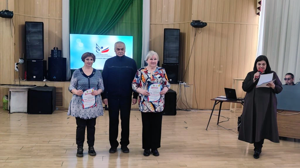 В центре стоят три человека, две женщины держат дипломы или грамоты. Справа женщина в длинном платье читает текст в микрофон.