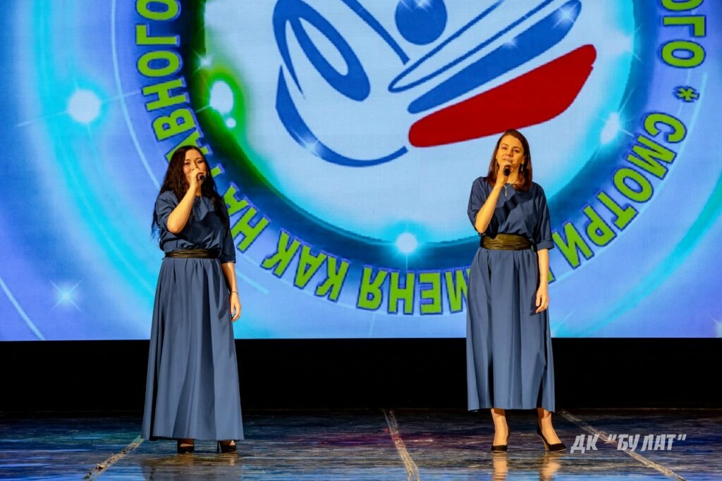 Две женщины поют на сцене с микрофонами, обе в длинных синих платьях с тёмными поясами; на экране сзади логотип и надпись по кругу