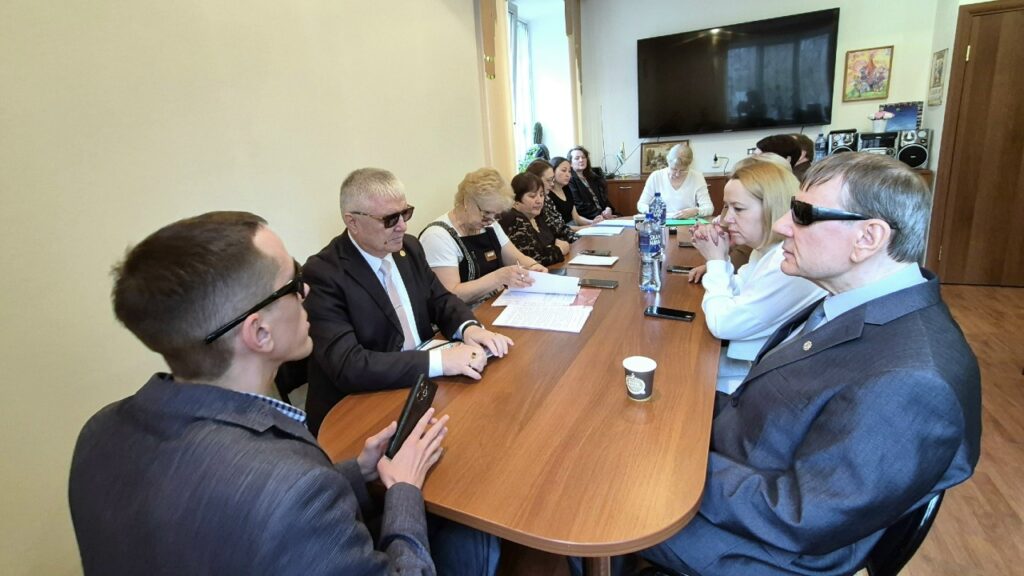 В офисе за длинным столом сидит группа людей на совещании; на столе бумаги, телефоны, бутылка воды и стаканчик, на стене большой телевизор.