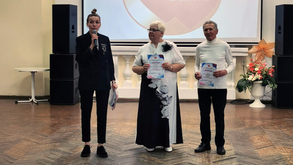 На сцене три человека. Слева ведущая в чёрном с микрофоном. В центре женщина в бело-чёрном платье с цветочным рисунком держит диплом. Справа мужчина в белой водолазке тоже держит диплом; сзади экран и большие колонки.