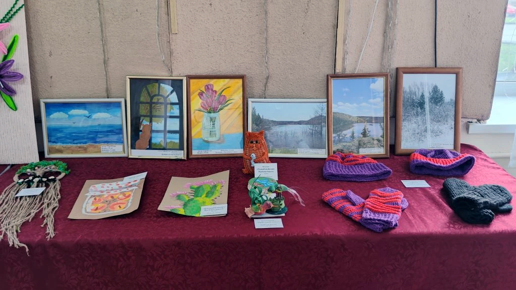 На столе выставка работ. Сзади в рамках несколько рисунков и фотографий: море, окно, цветы в вазе, пейзажи, зимний лес. Впереди лежат поделки: вязаные шапки и варежки ярких цветов, плетёные и бумажные работы, маленькие фигурки. Всё аккуратно разложено на бордовой скатерти.