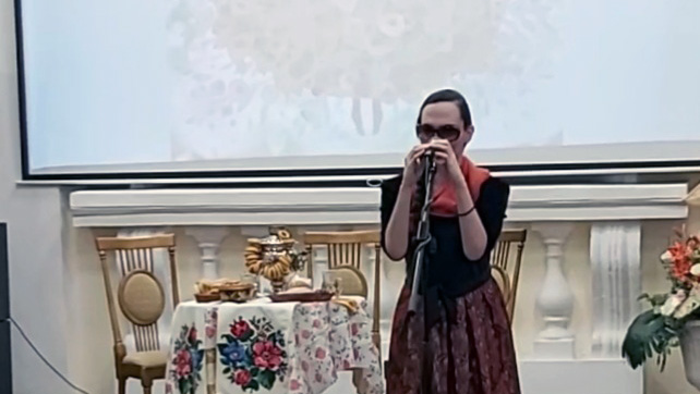 Женщина в тёмных очках стоит у микрофона и поёт. На ней чёрный верх, красный платок на шее и тёмная юбка с узором. Слева стол с цветочной скатертью и предметами, рядом два стула; сзади большой экран.