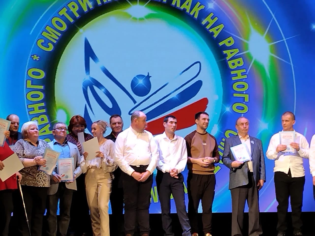 Группа людей стоит на сцене, многие держат дипломы/грамоты; на большом экране позади круглый логотип и надпись по кругу