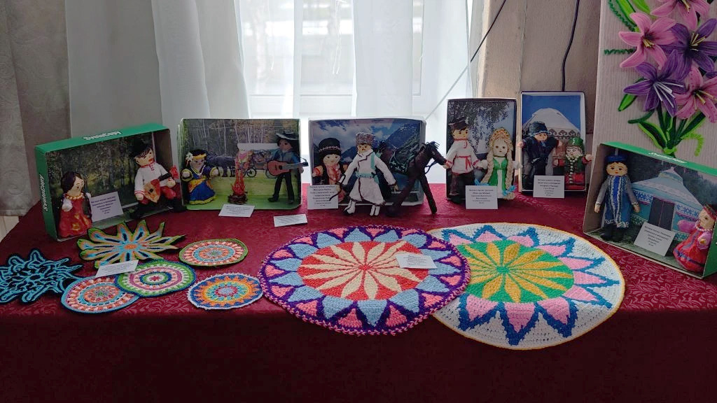 На столе выставка.
Сзади стоят композиции с куклами в национальных костюмах, каждая в своей коробке‑сценке.
Спереди разложены яркие вязаные и плетёные круглые салфетки и узорные коврики с геометрическими орнаментами.