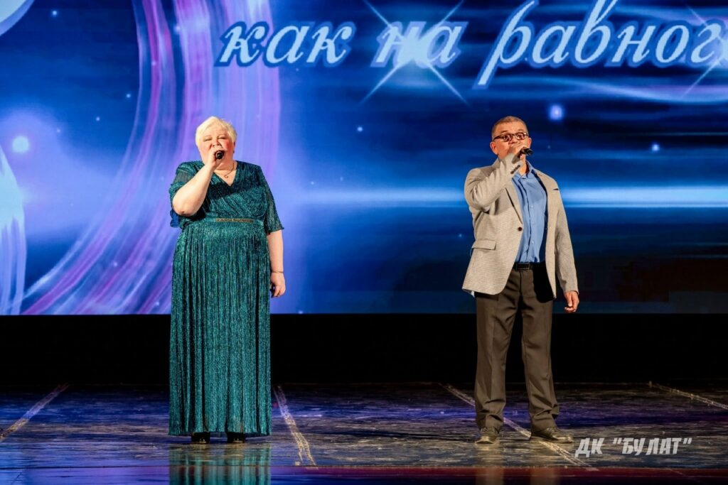 Два человека поют на сцене с микрофонами: женщина в длинном зелёном блестящем платье и мужчина в очках в сером пиджаке; на фоне экран с надписью