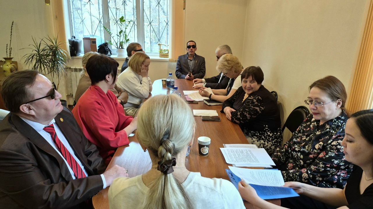 В офисе за длинным столом идёт совещание: несколько мужчин и женщин сидят вокруг, на столе бумаги, папки и бутылка воды; у окна в конце стола сидит мужчина в тёмных очках.