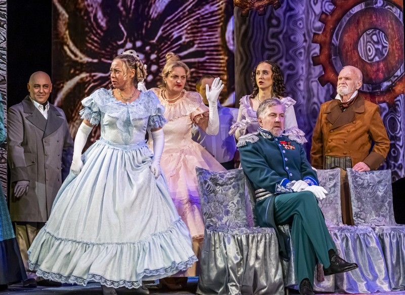Театральная сцена: несколько актёров в исторических костюмах; женщина в пышном голубом платье в центре, мужчина в зелёной форме с медалями сидит в кресле.