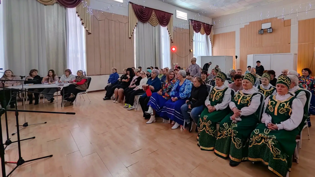 рительный зал. Люди сидят рядами на стульях. Справа целая группа женщин в одинаковых зелёных народных костюмах с золотым узором и кокошниками. В центре и слева зрители в обычной одежде, ближе к левому краю за столом сидит несколько женщин, похожих на жюри или организаторов. На переднем плане видны стойки с микрофонами.