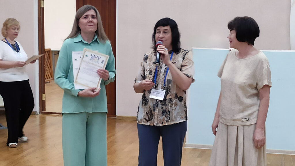 Три женщины в зале: одна в светло-зелёном костюме с дипломами, вторая с микрофоном и бейджем, третья в светлом платье рядом; на заднем плане ещё одна женщина с бумагами у двери.