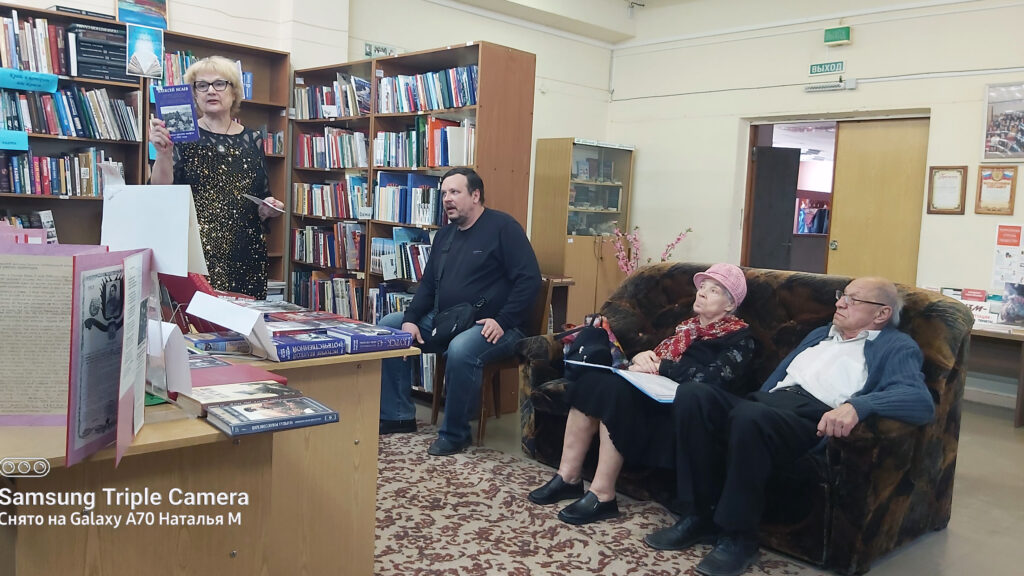 Женщина в библиотеке стоит у стола с разложенными книгами и показывает книгу.
Перед ней трое слушателей: мужчина на стуле и пожилая пара на диване.