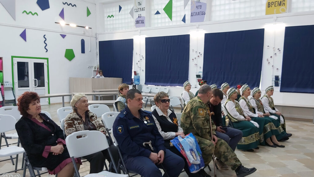 В зале на складных стульях сидят зрители, среди них несколько людей в форме и женщины в народных зелёно-золотых костюмах.
