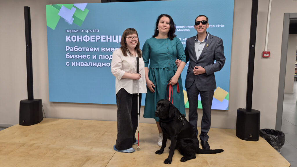 На сцене перед экраном с надписью о конференции «Работаем вместе: бизнес и люди с инвалидностью» стоят две женщины и мужчина, рядом чёрная собака‑поводырь.