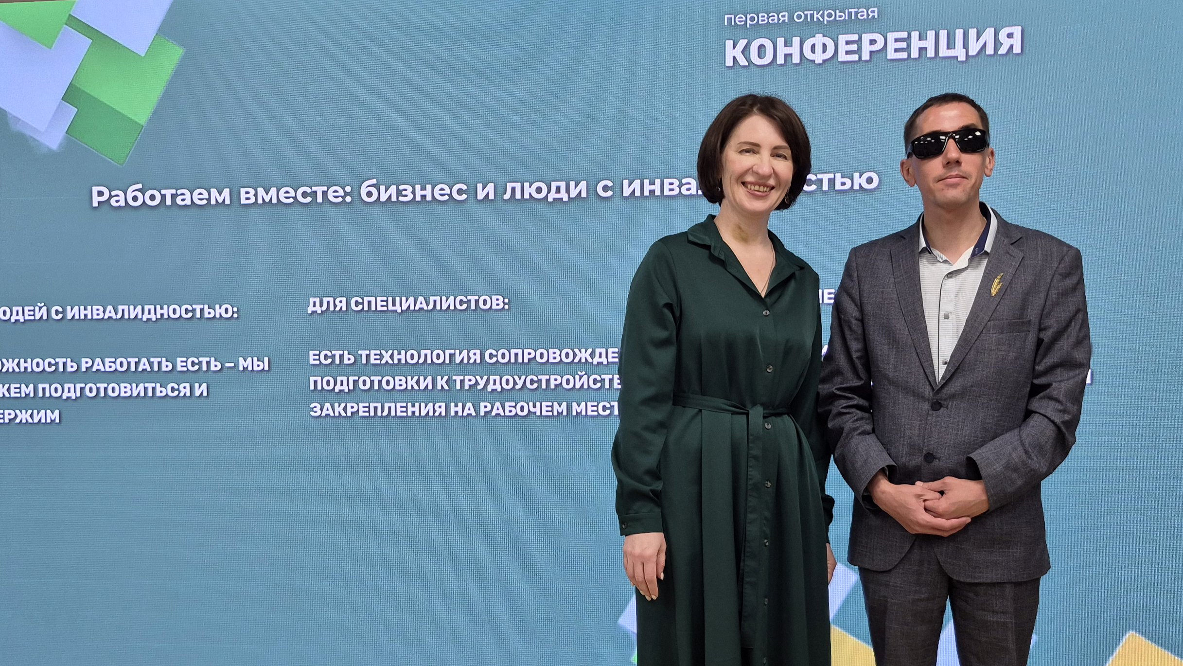 На фоне экрана с надписью «Работаем вместе: бизнес и люди с инвалидностью» стоят двое людей. Слева женщина в длинном тёмно‑зелёном платье, справа мужчина в сером костюме и тёмных очках.