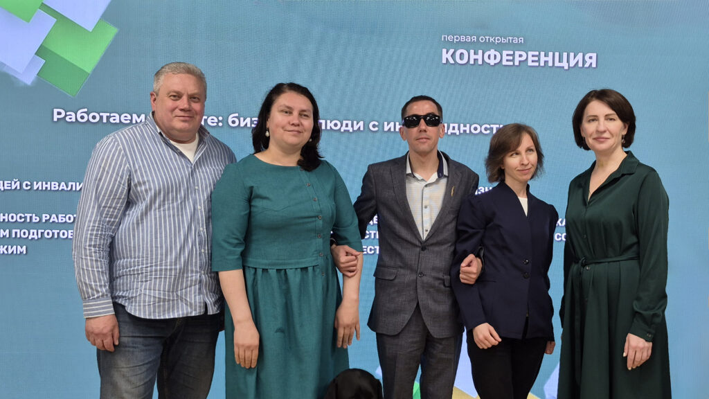 На фоне экрана с надписью о конференции стоит группа из пяти человек.