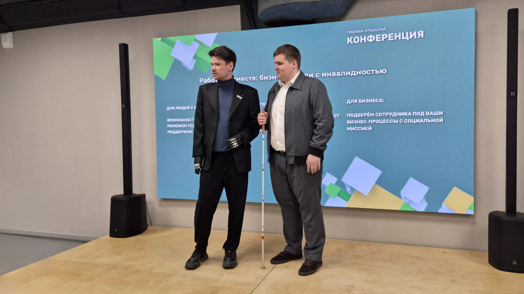 Двое мужчин стоят на сцене на фоне экрана с надписью о конференции «Работаем вместе: бизнес и люди с инвалидностью».
Один мужчина в чёрном костюме с бионическими протезами кистей, второй в сером костюме держит белую трость.