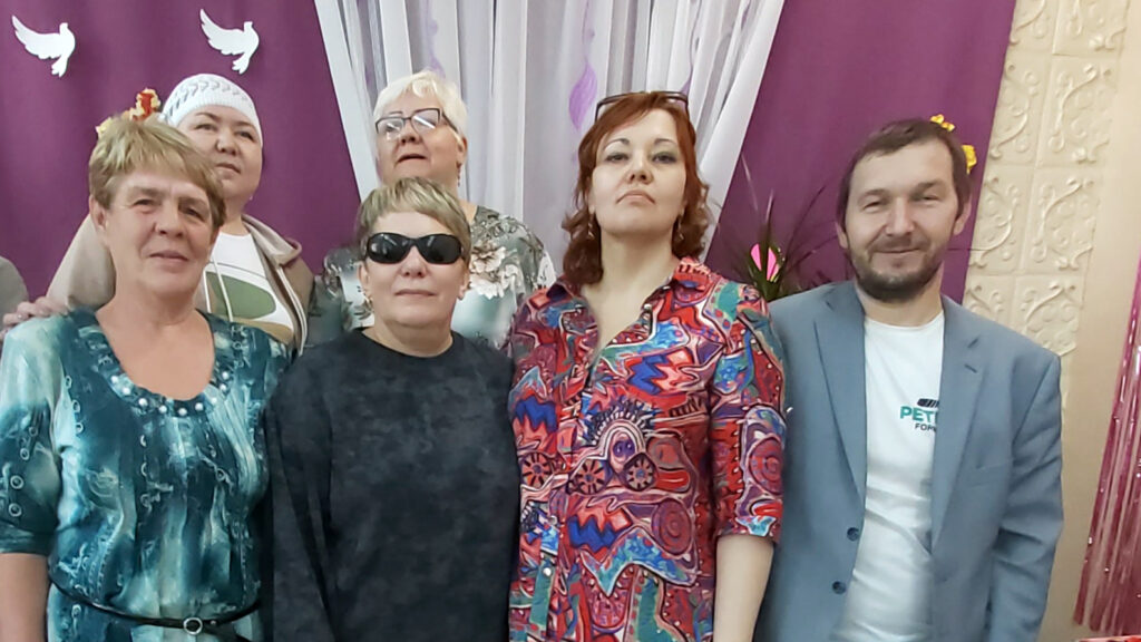 Шесть взрослых позируют в помещении на фоне фиолетового занавеса с белыми птичками; стоят плотной группой, смотрят в камеру.