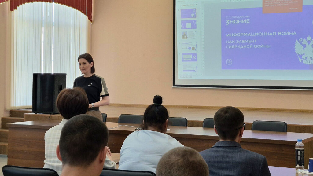Женщина проводит лекцию в аудитории перед сидящими людьми, на экране слайд презентации с заголовком «Информационная война как элемент гибридной войны».