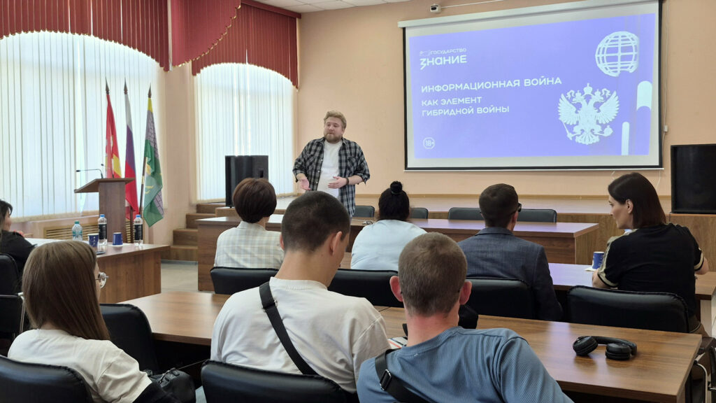 Мужчина выступает с лекцией перед сидящими слушателями; на экране слайд «Информационная война как элемент гибридной войны».