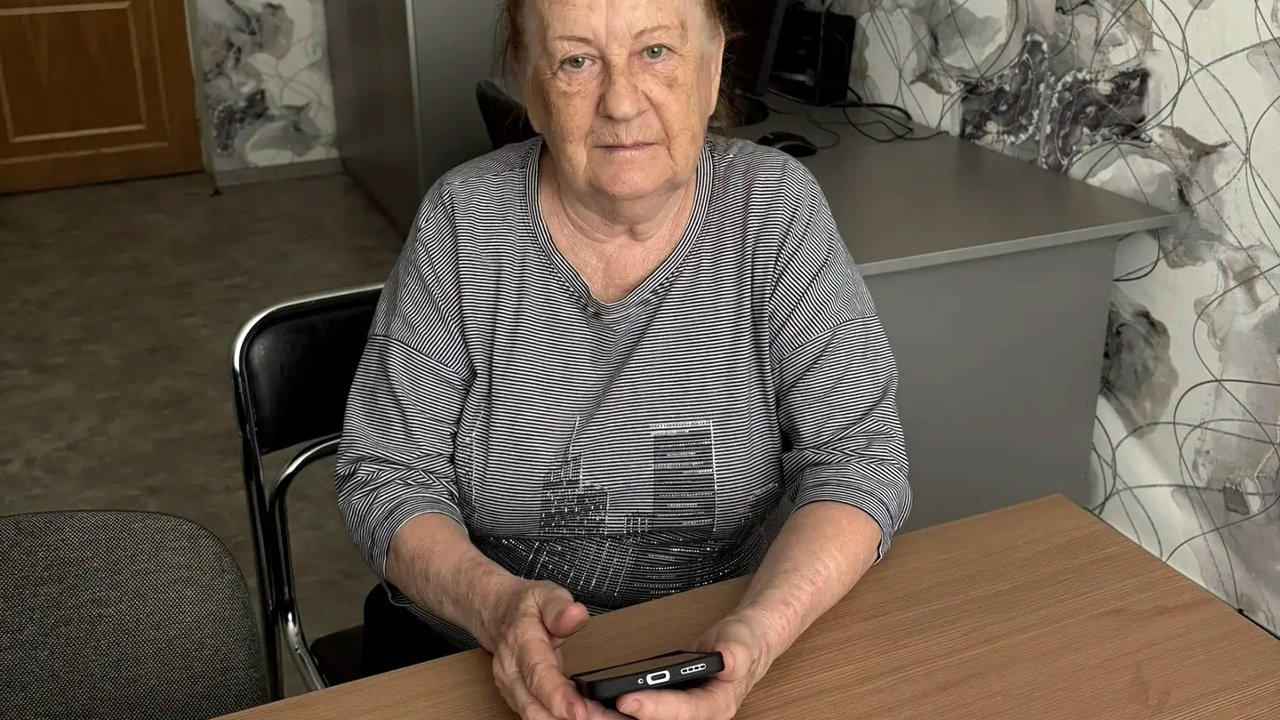 Пожилая женщина сидит за столом в комнате и держит в руках небольшое чёрное электронное устройство. На ней серая полосатая футболка с рисунком домов. На заднем плане стол с техникой и стул.