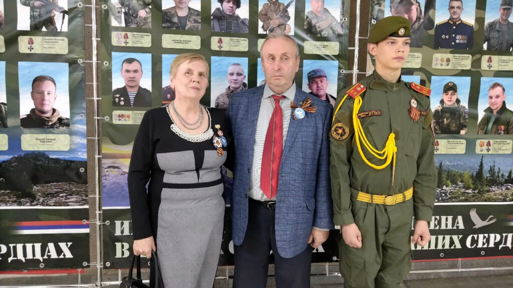 На фото трое людей на фоне стенда с фотографиями военных.
Слева женщина в серо-чёрном платье с медалями, в центре мужчина в пиджаке и красном галстуке с георгиевской ленточкой, справа юноша в зелёной военной форме с погонами и медалями.