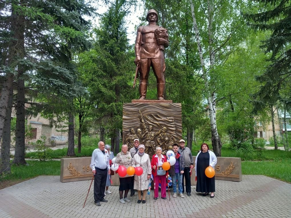 Группа людей с воздушными шарами стоит у большого бронзового памятника солдату в парке.