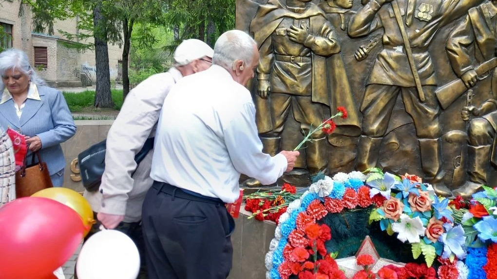 Пожилые люди у бронзового памятника солдатам возлагают красные гвоздики и венки, рядом видны цветные шары.