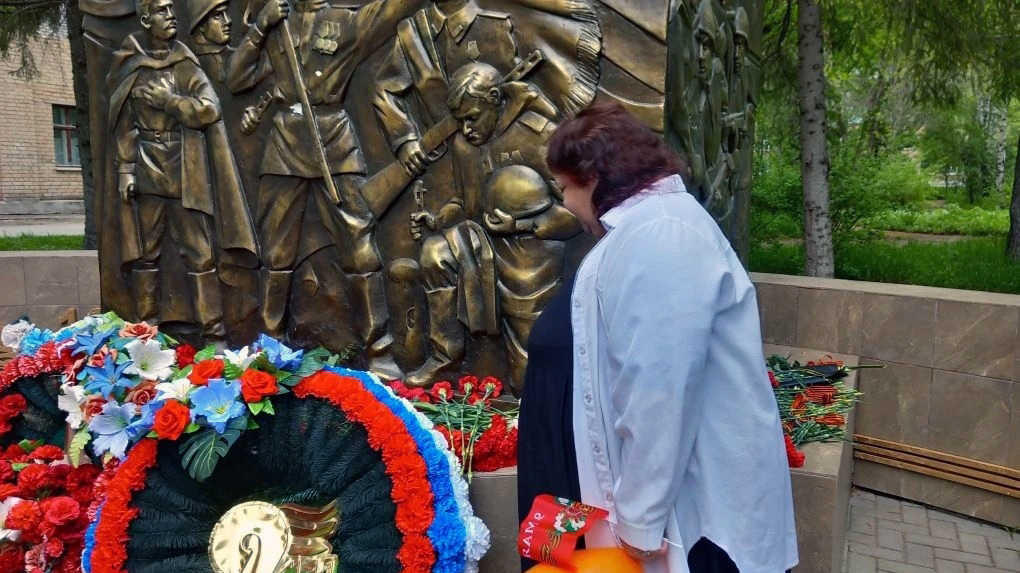 Женщина в светлой куртке стоит у бронзового памятника солдатам, перед ним лежат красные гвоздики и пышные венки из разноцветных цветов.