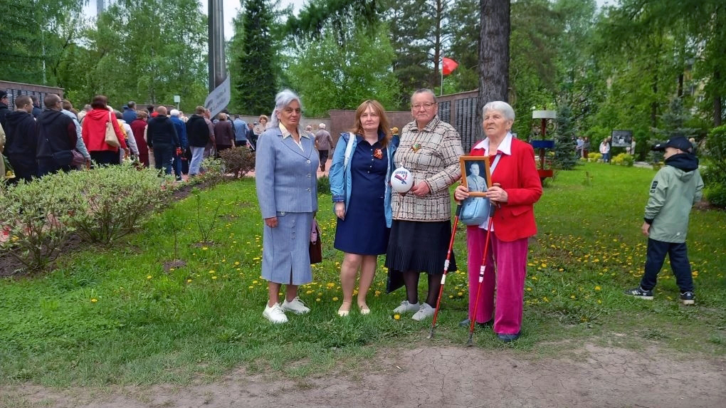 Четыре женщины стоят в парке у мемориала, одна держит фотографию в рамке, другая — белый шар; вокруг много людей и зелёных деревьев.