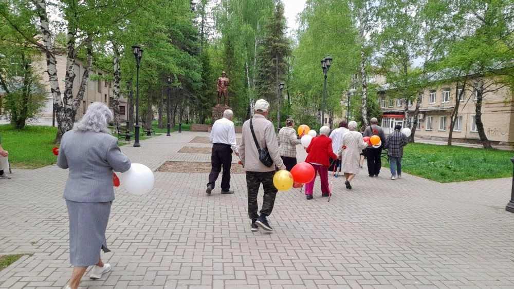 Группа людей с воздушными шарами идёт по аллее в сторону памятника солдату в парке.