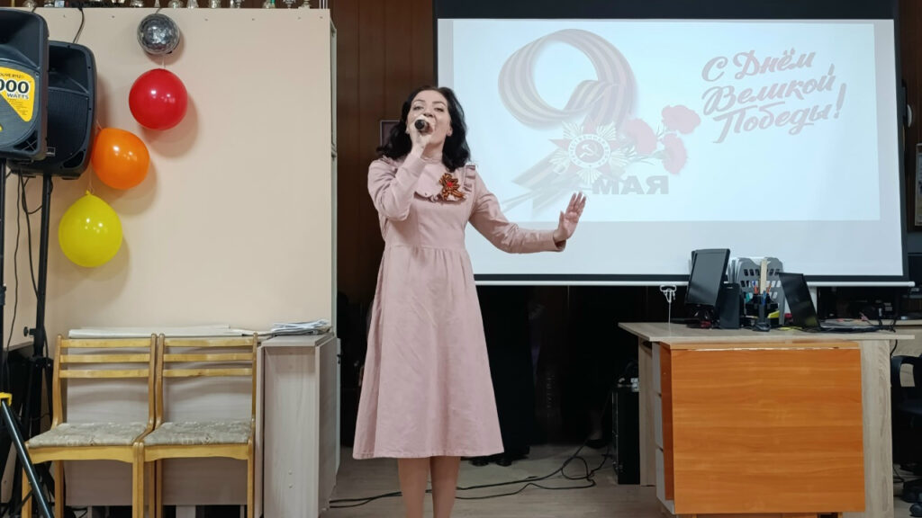 На фото женщина в розовом платье поёт в микрофон перед экраном.
На экране надпись: «С Днём Великой Победы! 9 Мая».
Слева висят три воздушных шара, справа стоит стол с компьютерами.