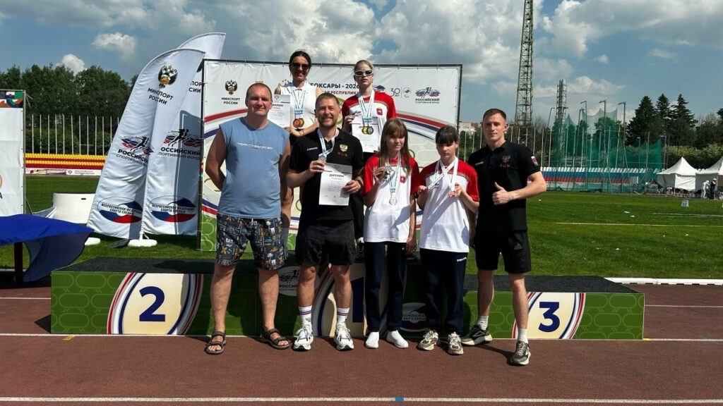 Группа спортсменов и тренеров стоит на фоне пьедестала и баннера «Чебоксары – 2026» на стадионе; несколько человек с медалями и дипломами, справа мужчина показывает жест «класс».