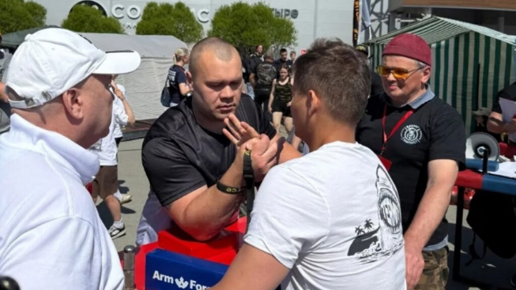 На фото соревнование по армрестлингу на улице.
Двое мужчин напротив друг друга готовятся бороться за столом для армрестлинга, судья в кепке и чёрной футболке наблюдает рядом, ещё один мужчина в белой одежде стоит сбоку.