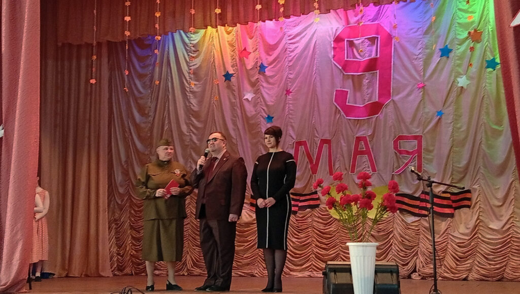 На сцене под надписью «9 МАЯ» стоят трое выступающих (женщина в военной форме, мужчина с микрофоном, женщина в чёрном платье), перед ними вазон с красными гвоздиками.
