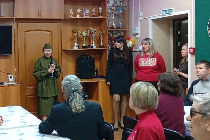 у двери стоят три женщины, одна в синнем пиджаке говорит, рядом мужчина сидит и играет на аккордеоне, участники слушают за столами.