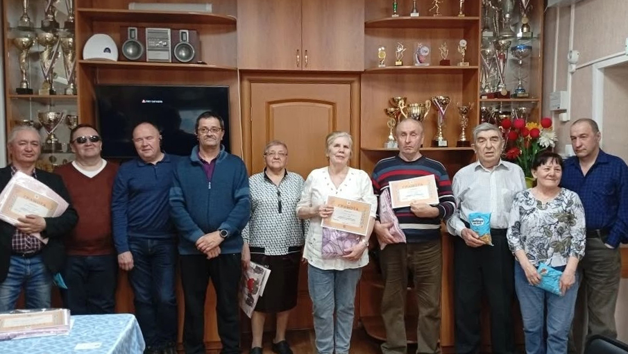 На фото группа из девяти людей стоит в ряд в комнате с витринами кубков. Несколько человек в центре держат в руках грамоты и подарки, остальные тоже с пакетами или коробками,