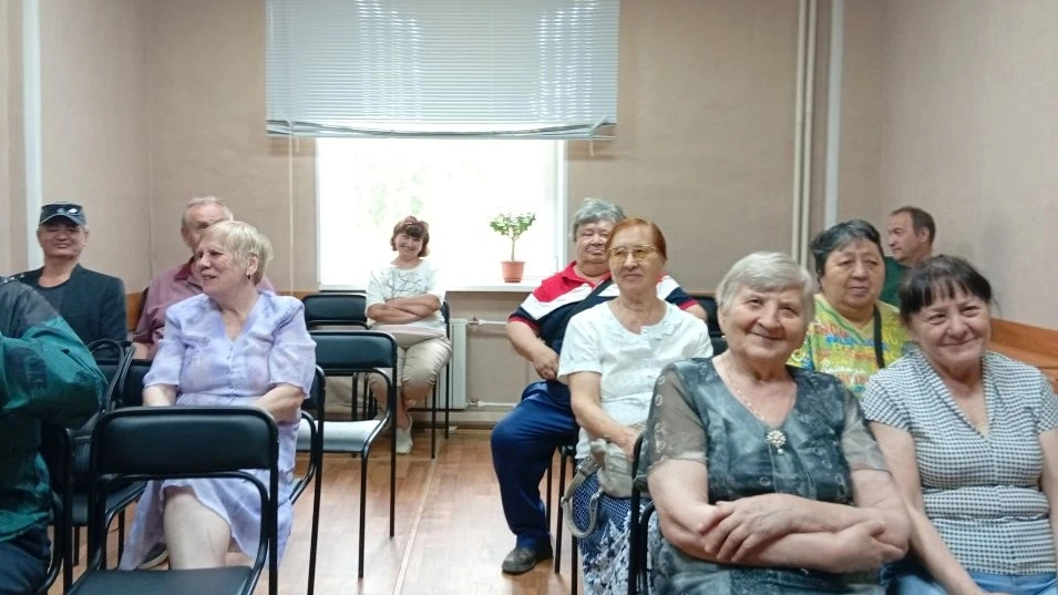 Группа пожилых людей сидит рядами на стульях в небольшой комнате, многие улыбаются; на заднем плане окно с опущенной шторой и цветок в горшке на подоконнике.