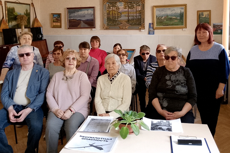 Групповое фото людей в комнате, многие пожилые, некоторые в тёмных очках. На столе перед ними лежат бумаги с заголовком «БЕСПИЛОТНАЯ ОПАСНОСТЬ», на стенах висят пейзажные картины.