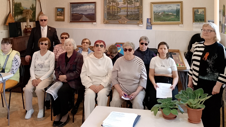 Группа людей в комнате, в основном пожилые женщины, многие в тёмных очках, сидят и стоят для общего фото.
Справа стоит женщина в чёрно‑белом свитере с георгиевской ленточкой, слева и на столе видны горшки с зелёными растениями, на стене сзади висят пейзажные картины.
