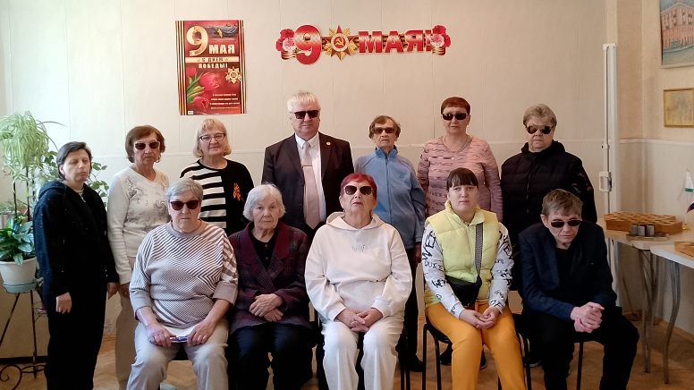 Групповое фото в комнате: один мужчина и несколько женщин, большинство в тёмных очках, на фоне надписи и плаката «9 Мая».
