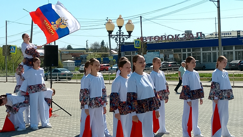 Группа девушек в белых костюмах выступает на площади, слева построена живая пирамида, на вершине которой девушка размахивает российским флагом; на заднем плане здание с надписью «АВТОВОКЗАЛ».