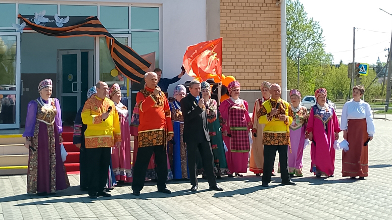 Группа людей в ярких народных костюмах поёт у входа в здание, позади развёрнуты красный флаг и большая георгиевская лента.